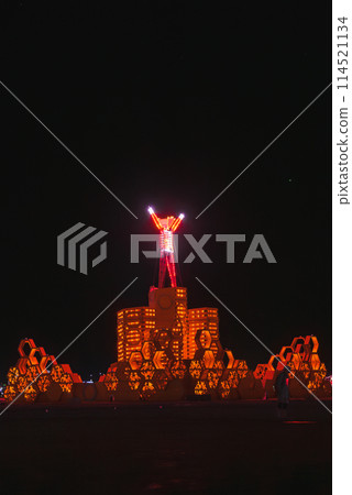 Nighttime scene at desert based music festival with large illuminated humanoid figure, glowing platform, and temporary cityscape against dark desert backdrop. 114521134