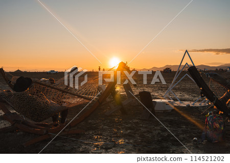 Scenic desert sunset with hammocks at a music festival location. Tranquil setting with golden sky, distant mountains, and festival infrastructure in background. 114521202