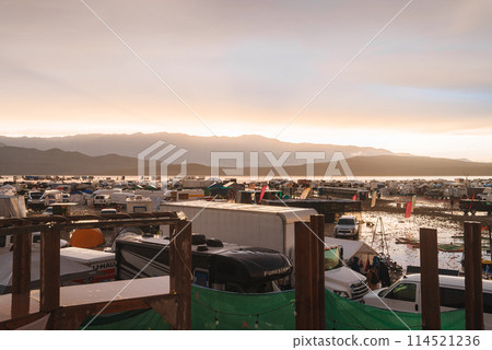 Desert music and art festival scene from an elevated view at either sunrise or sunset. Vehicles and creative structures against a backdrop of a desert landscape. 114521236