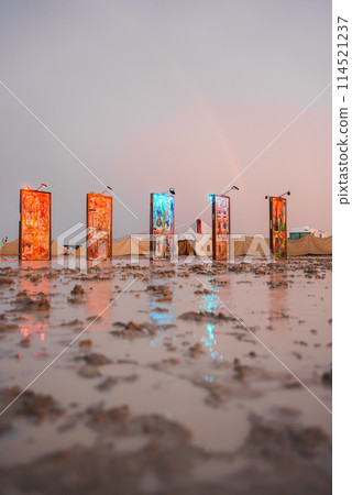 Colorful panels with unique artwork stand in a desert after rain, reflecting in puddles. Soft sky hues add to the surreal, serene atmosphere. Potentially part of a festival. 114521237