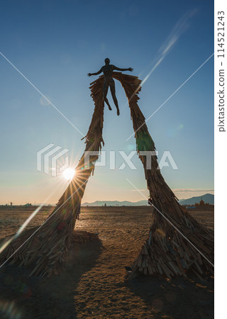 Striking art installation in desert setting under clear blue skies features two wooden structures resembling a portal with figure at the top at golden hour. 114521243