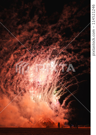 Vibrant fireworks illuminate night sky at music festival in desert setting, bursting pink, white, and green hues. Silhouettes of attendees marvel below. 114521246