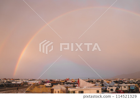 A music and art festival hosted in a desert, featuring a vibrant community living in tents and makeshift shelters, under a twilight sky with a rainbow. 114521329