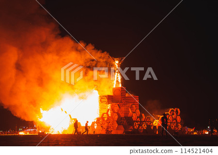 Large wooden structure ablaze at desert festival, intricate design with geometric patterns, silhouettes of onlookers. Location hint Burning Man style event. Large wooden structure ablaze at desert festival, intricate design with geometric patterns, silhouettes of onlookers. Location hint Burning Man style event. 114521404