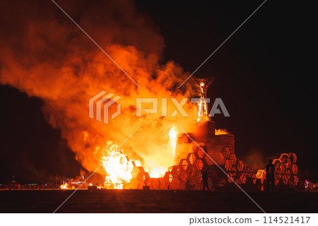 Dramatic night scene with burning wooden structure at desert art festival. Symbolizes impermanence, creativity, and community. Lights and people in background. Dramatic night scene with burning wooden structure at desert art festival. Symbolizes impermanence, creativity, and community. Lights and people in background. 114521417
