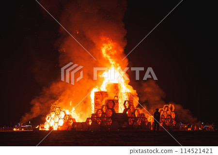 Fiery blaze consumes wooden structure in desert festival setting at night, creating bright orange flames illuminating the dark sky in a remote location. 114521421