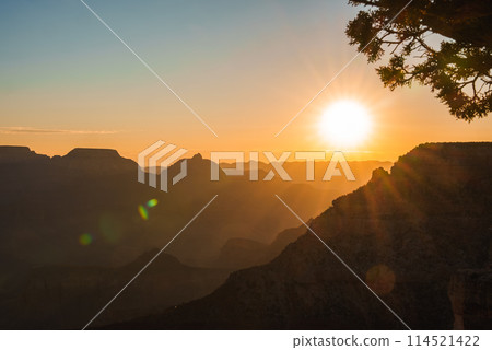 Sunset landscape with rugged mountains fading into distance, warm golden glow in the sky. Silhouetted tree branch frames the scene, lens flares add interest. Peaceful, majestic mood. 114521422