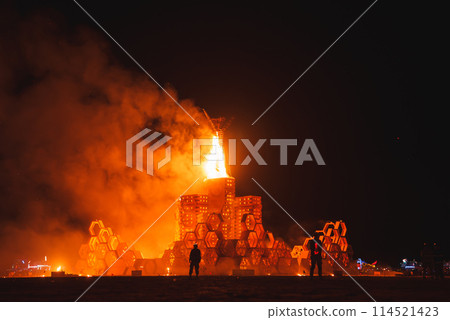 Night scene at desert music and art festival. Large, intricate structure engulfed in flames, people watching in awe. Dark desert horizon in background. 114521423