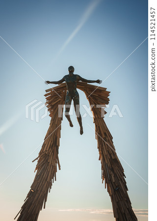 Person floating mid air in front of wooden wing like structure at desert music festival. Symbolic flight captured against clear blue sky. Artistic, immersive experience. 114521457