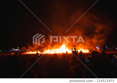 Nighttime desert festival scene with a crowd silhouetted against intense flames. Warm orange glow, structures in the background, isolated desert location. Nighttime desert festival scene with a crowd silhouetted against intense flames. Warm orange glow, structures in the background, isolated desert location. 114521473