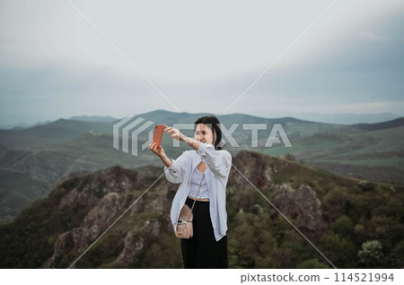 Happy woman tourist hiking up mountain enjoying nature. Landscape in cloudy gloomy windy weather. Smiling female in white shirt takes selfie pictures on smartphone, enjoying travel. Freedom concept. Happy woman tourist hiking up mountain enjoying nature. Landscape in cloudy gloomy windy weather. Smiling female in white shirt takes selfie pictures on smartphone, enjoying travel. Freedom concept. 114521994