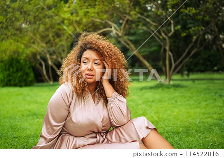 Close up portrait of Young beautiful African American woman enjoys a walk in summer public green park. A happy woman plus size wearing silk pink dress sitting resting on grass. 114522016