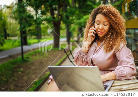 Attractive happy stylish plus size African American Black business woman speaks phone working online on laptop computer at sidewalk cafe workspace outdoor. Remote work, distance education. 114522017