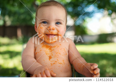 Cute adorable infant funny dirty baby trying to eat himself with fruit or vegetable puree smeared on his face. Baby's first complementary solid food. Organic natural ingredients. Summer outdoor. 114522018