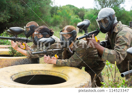 Excited male and female paintball players in protective uniform aiming and shooting with guns outdoor 114523347