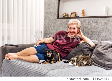 Mature russian woman smiling and sitting on the sofa with her pets cat and dog, drinking wine and looking at the camera Mature russian woman smiling and sitting on the sofa with her pets cat and dog, drinking wine and looking at the camera 114523617