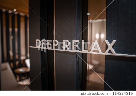 Glass door with DEEP RELAX in modern font stands out against dark frame. Through the door, a serene spa setting in luxury Zermatt, Switzerland hotel. 114524519