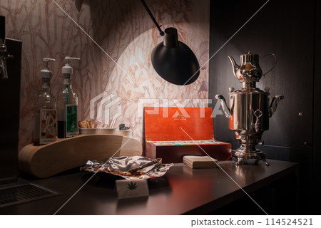 Luxury hotel countertop in Zermatt ski resort, Switzerland. Dark material contrasts with transparent bottles, white cups, orange box, and silver samovar. Modern lamp provides warm light ambiance. 114524521