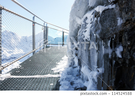 Metal walkway in snowy mountainous Murren, Switzerland. Chain link fence, icy rock face, snow on ground, sunny day, mountains in background. Tranquil isolation. 114524588