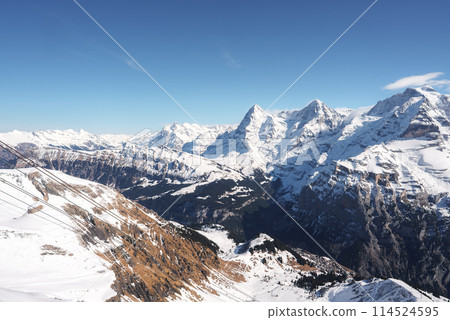 Breathtaking view of Murren ski resort in Switzerland. Snow covered slopes, rocky terrain, ski lift system, deep valley, Swiss Alps peaks, clear blue sky. 114524595