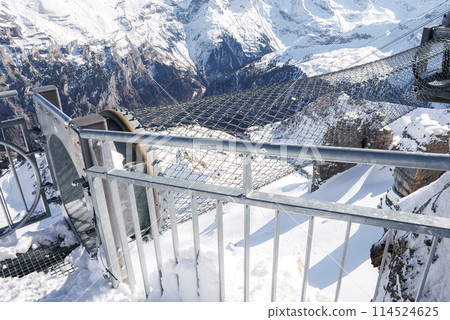 Metal viewing platform at Murren ski resort, Switzerland, offers panoramic snowy mountain views under clear skies. Sturdy railing and mesh floor ensure safety. Tranquil, untouched nature. 114524625