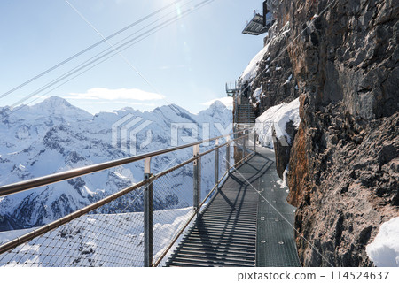 Metal walkway on rocky cliff in Murren ski resort, Switzerland. Mesh structure, railing, snow covered peaks, blue sky. Typical winter setting for skiers. Metal walkway on rocky cliff in Murren ski resort, Switzerland. Mesh structure, railing, snow covered peaks, blue sky. Typical winter setting for skiers. 114524637