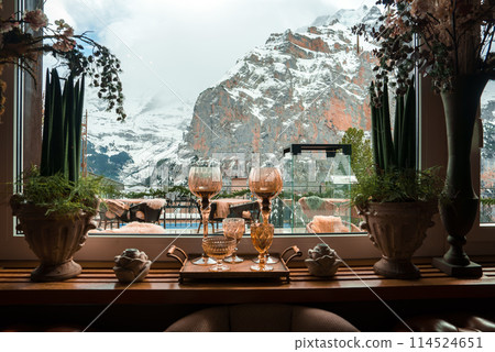 Cozy interior scene with snowy mountain landscape view through window. Elegant glassware on wooden table, decorative tea set, potted plants. Comfort and luxury vibes. High altitude or alpine setting. 114524651