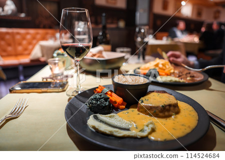Dining scene with gourmet steak and vegetables on plate, glass of red wine, and cozy ambiance. Smartphone and fork on table. Other diners in background. 114524684