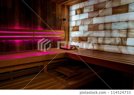 Interior of sauna room featuring wood panels, salt bricks on wall. Wooden benches for seating arranged for heat circulation. Pink lighting creates tranquil ambiance. Interior of sauna room featuring wood panels, salt bricks on wall. Wooden benches for seating arranged for heat circulation. Pink lighting creates tranquil ambiance. 114524733