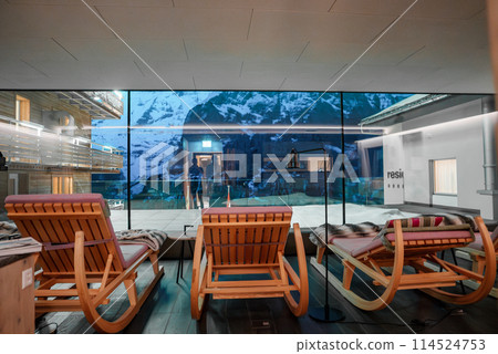 Modern indoor relaxation space with wooden flooring, orange lounge chairs, snowy mountain view through a glass window, and alpine ambiance. Luxurious and serene. Modern indoor relaxation space with wooden flooring, orange lounge chairs, snowy mountain view through a glass window, and alpine ambiance. Luxurious and serene. 114524753