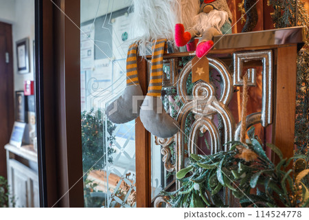 Whimsical holiday gnome climbing gold railing with pine branch decorations in festive indoor setting with blurry reception area visible through window. Whimsical holiday gnome climbing gold railing with pine branch decorations in festive indoor setting with blurry reception area visible through window. 114524778