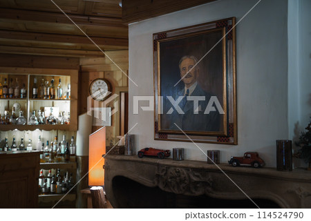 Interior scene in wood paneled room with a portrait of a man in a suit and tie. Stone fireplace mantel, cabinet with bottles, clock on the wall, warm lighting. 114524790