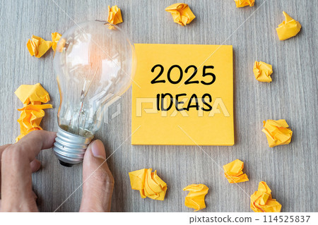 2025 Idea words on yellow note and crumbled paper with Businessman holding lightbulb on wooden table background. New Year New Start Creative, Innovation, Imagination, Resolution and Goal concept 114525837