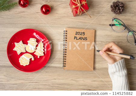 Woman hand writing 2025 PLANS on notebook with Christmas cookies on table. Xmas, Happy New Year, Goals, Resolution, To do list, and Strategy concept 114525882