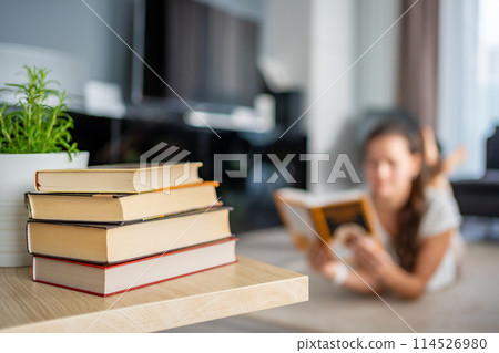Stack of books in the foreground and woman reads a book lying down in the background Stack of books in the foreground and woman reads a book lying down in the background 114526980