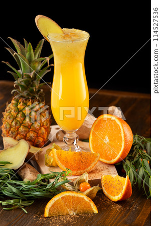 Closeup glass of fresh orange and pineapple juice decorated with ginger and fruits on a wooden table isolated at black background. Closeup glass of fresh orange and pineapple juice decorated with ginger and fruits on a wooden table isolated at black background. 114527536