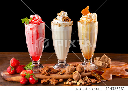 Three sweet milkshakes with nuts, caramel, strawberry and whipped cream at a wooden board on table background. Three sweet milkshakes with nuts, caramel, strawberry and whipped cream at a wooden board on table background. 114527581