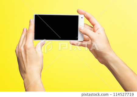 Mockup of female hands holding white frameless cellphone with black screen and making selfie at isolated yellow background. 114527719