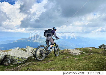 Cyclist man riding electric mountain bike outdoors. Male tourist biking along grassy trail in the mountains, wearing helmet and backpack. Concept of sport, active leisure and nature. Cyclist man riding electric mountain bike outdoors. Male tourist biking along grassy trail in the mountains, wearing helmet and backpack. Concept of sport, active leisure and nature. 114529919