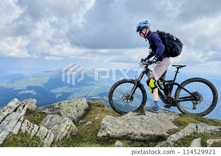 Cyclist man riding electric mountain bike outdoors. Male tourist biking along grassy trail in the mountains, wearing helmet and backpack. Concept of sport, active leisure and nature. Cyclist man riding electric mountain bike outdoors. Male tourist biking along grassy trail in the mountains, wearing helmet and backpack. Concept of sport, active leisure and nature. 114529921