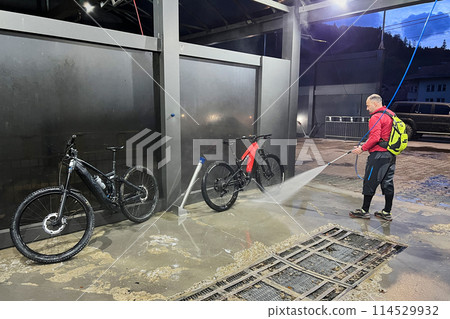Cyclist using high-pressure water spray to clean two electric mountain bikes on outdoor washing station. Cyclist using high-pressure water spray to clean two electric mountain bikes on outdoor washing station. 114529932
