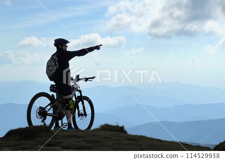 Cyclist man riding electric bike outdoors. Silhouette of male tourist resting on top of hill, pointing on beautiful mountain landscape, wearing helmet and backpack. Concept of active leisure. Cyclist man riding electric bike outdoors. Silhouette of male tourist resting on top of hill, pointing on beautiful mountain landscape, wearing helmet and backpack. Concept of active leisure. 114529938