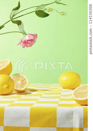 A table covered by a yellow checkered tablecloth containing some fresh yellow lemon above on light green background, a pink eustoma branch decorated. Blank space on table for showing product A table covered by a yellow checkered tablecloth containing some fresh yellow lemon above on light green background, a pink eustoma branch decorated. Blank space on table for showing product 114530308