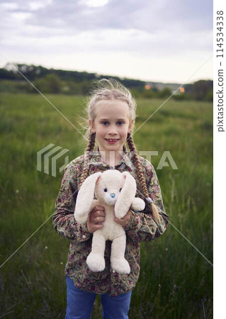 a beautiful blond girl with her toy bunny is playing in the field a beautiful blond girl with her toy bunny is playing in the field 114534338