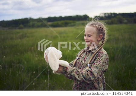 a beautiful blond girl with her toy bunny is playing in the field 114534341