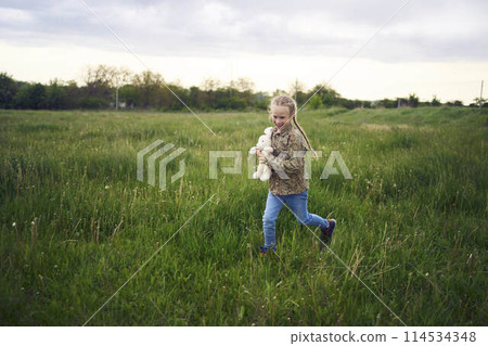 a beautiful blond girl with her toy bunny is playing in the field 114534348