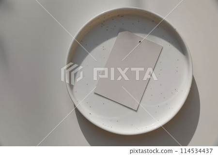Blank gift tag label mockup in sunlight. Spotted ceramic plate on beige table background. Soft shadows. Neutral wedding stationery. Food, restaurant, craft concept. Flat lay. Top view. 114534437