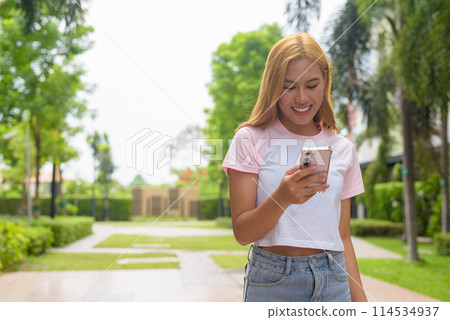 Portrait of beautiful Asian girl with blonde hair smiling using phone outdoors Portrait of beautiful Asian girl with blonde hair smiling using phone outdoors 114534937