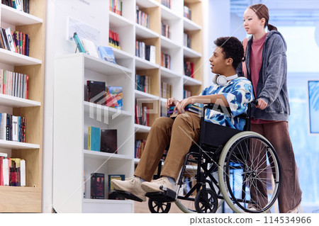 Full length portrait of Middle Eastern boy using wheelchair choosing books on shelves in school library with friend assisting copy space 114534966