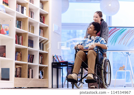 Full length portrait of teenage boy with disability choosing books in school library with smiling girl assisting copy space 114534967